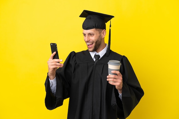 Jovem universitário, homem branco, isolado em um fundo amarelo, segurando um café para levar e um celular
