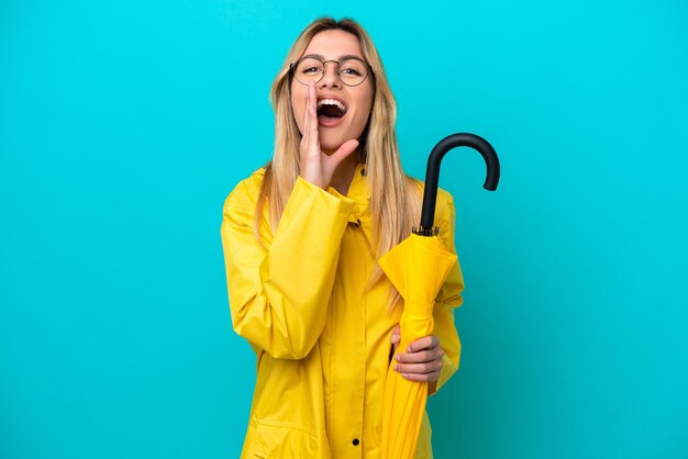 Jovem uruguaia com casaco à prova de chuva e guarda-chuva isolado em fundo azul gritando com a boca aberta