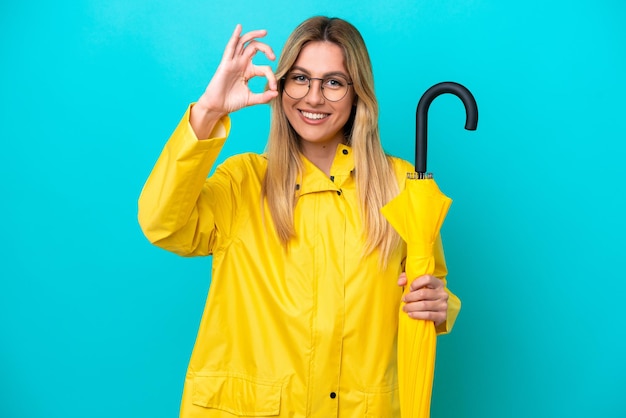 Jovem uruguaia com casaco à prova de chuva e guarda-chuva isolado em fundo azul, mostrando sinal de ok com os dedos