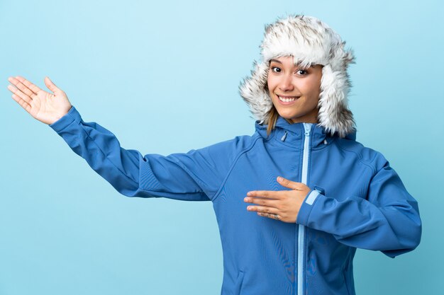 Jovem uruguaia com chapéu de inverno na parede azul, estendendo as mãos para o lado para convidar para vir