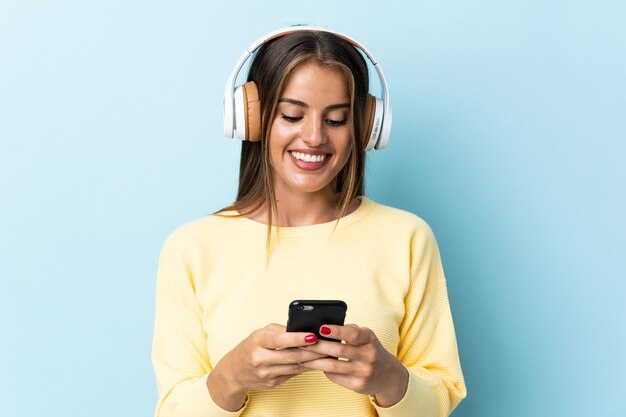 Jovem uruguaia isolada em um fundo azul ouvindo música e olhando para o celular