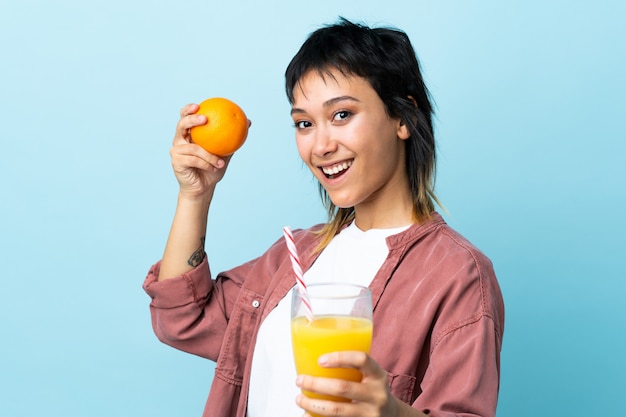 Jovem uruguaia sobre fundo azul isolado, segurando uma laranja e um suco de laranja