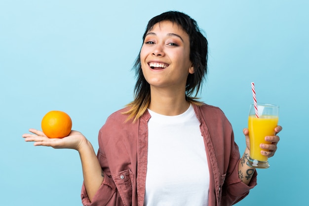 Jovem uruguaia sobre fundo azul isolado, segurando uma laranja e um suco de laranja