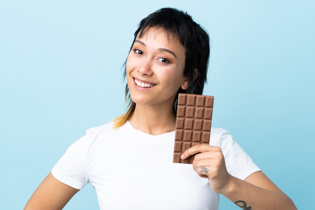 Jovem uruguaia sobre fundo azul isolado, tomando um comprimido de chocolate e feliz