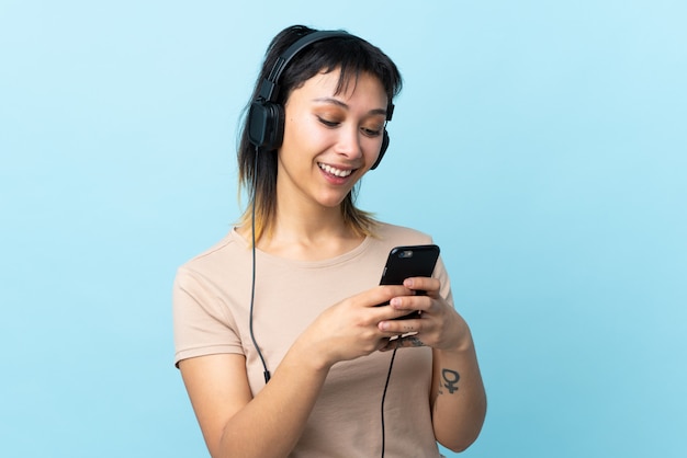 Jovem uruguaia sobre parede azul, ouvindo música e olhando para o celular