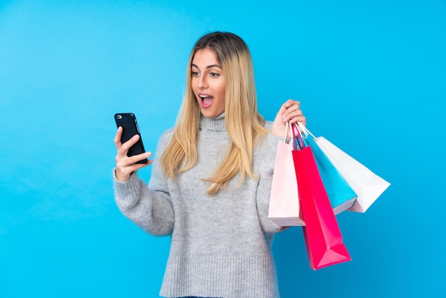 Jovem uruguaia sobre parede isolada parede azul segurando sacolas de compras e escrever uma mensagem com o celular para um amigo