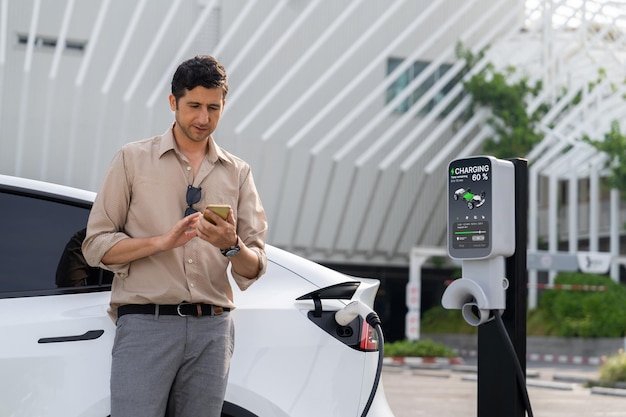 Jovem usa smartphone para pagar eletricidade em estação pública de carregamento de carros elétricos no estacionamento do shopping comercial da cidade Estilo de vida urbano moderno, ambiental e sustentável com veículo elétrico Expediente