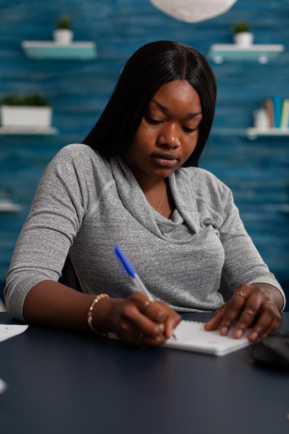 Jovem usando a caneta para escrever no arquivo do caderno enquanto trabalhava em casa na mesa. mulher escrevendo informações e fazendo anotações em papel de livro didático, fazendo trabalho remoto no projeto de negócios.