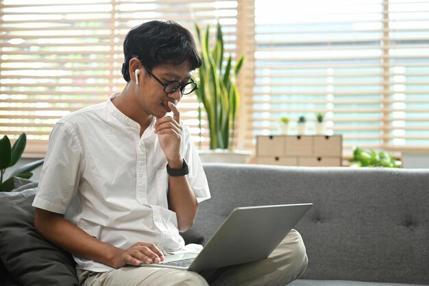 Jovem usando fone de ouvido sem fio navegando na internet com computador portátil na sala de estar brilhante