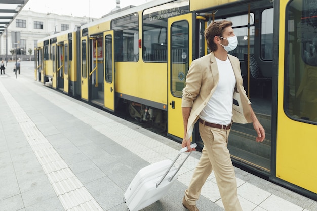 Jovem usando máscara médica protetora enquanto caminha na estação com uma mala branca