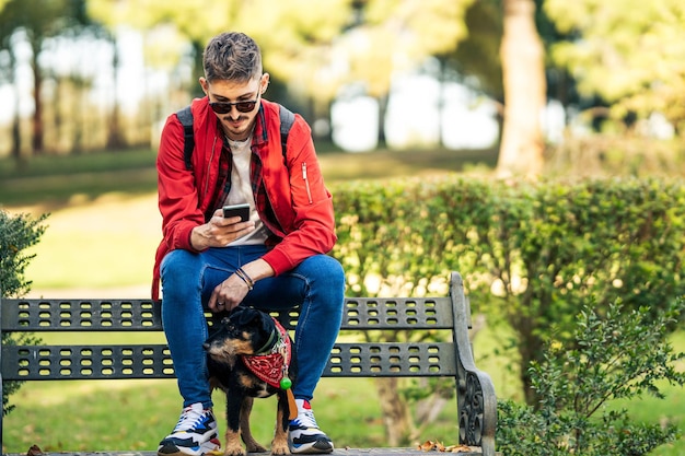 Jovem usando o celular enquanto está sentado em um banco de um parque com um cachorrinho entre as pernas
