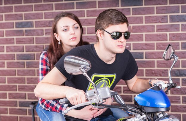 Jovem usando óculos escuros e mastigando palito de dente, andando de motocicleta em frente à parede de tijolos com uma jovem passageira nas costas