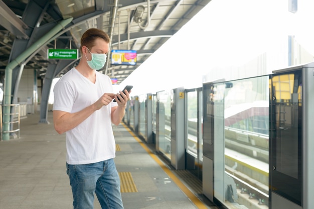 Foto jovem usando telefone e máscara para proteção contra surto do vírus corona na estação de trem