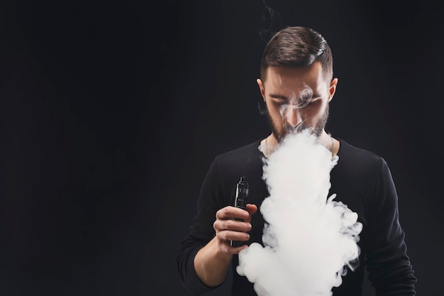 Foto jovem vaping, estúdio tiro. cara barbudo soprando uma nuvem de fumaça sobre fundo preto. conceito de fumo e vapor livre de nicotina, espaço de cópia.