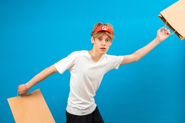 Jovem vendedor em camiseta branca casual com saco de papel correndo em fundo azul retrato de estúdio Conceito de estilo de vida de pessoas Mock up copy space Adolescente segurando saco de pacote com compras