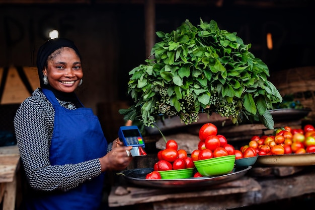 Jovem vendedor feliz do mercado africano segurando uma máquina de POS com um cartão ATM vendendo tomates