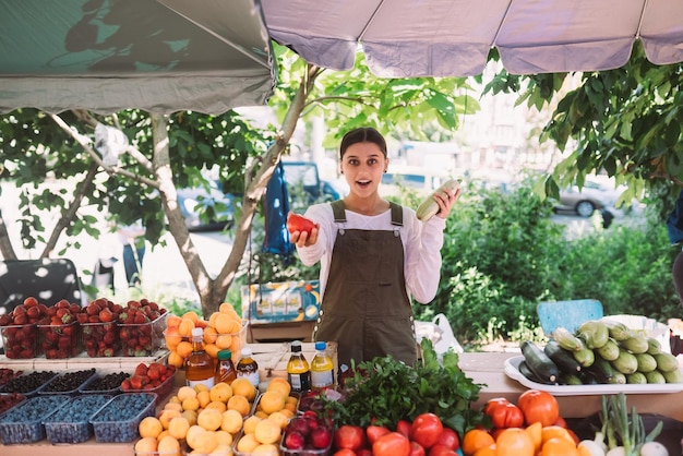 Jovem vendedora segurando abobrinha e tomate nas mãos