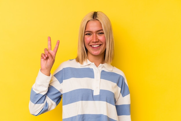 Jovem venezuelana isolada em fundo amarelo, mostrando o número dois com os dedos.