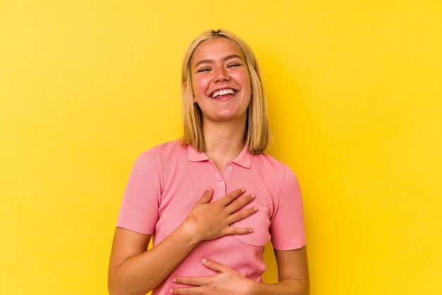 Jovem venezuelana isolada em fundo amarelo ri alegremente e se diverte mantendo as mãos na barriga.