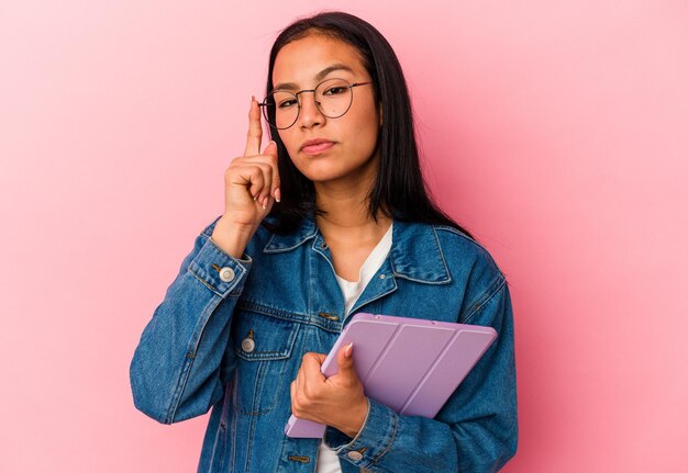 Jovem venezuelana segurando um tablet isolado no fundo rosa apontando o templo com o dedo pensando focado em uma tarefa