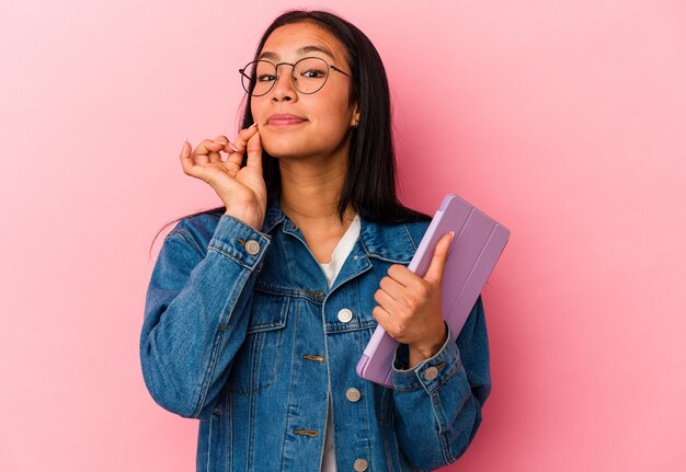 Jovem venezuelana segurando um tablet isolado no fundo rosa com os dedos nos lábios, mantendo um segredo