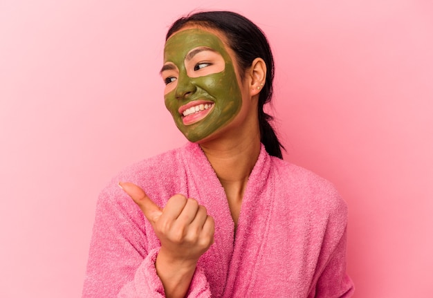 Jovem venezuelana vestindo um roupão de banho e máscara facial isolada em pontos de fundo rosa com o dedo polegar afastado, rindo e despreocupada.