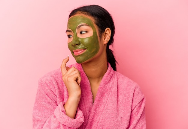 Jovem venezuelana vestindo um roupão de banho e máscara facial isolada em um fundo rosa relaxou pensando em algo olhando para um espaço de cópia.