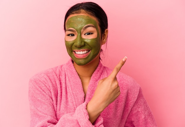 Jovem venezuelana vestindo um roupão de banho e máscara facial isolada na parede rosa sorrindo e apontando para o lado, mostrando algo no espaço em branco.