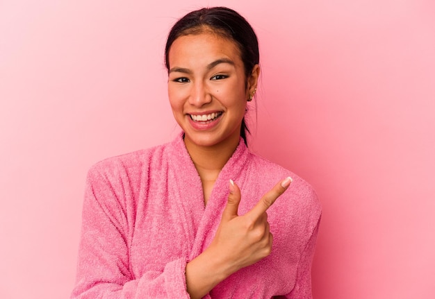 Jovem venezuelana vestindo um roupão de banho isolado no fundo rosa sorrindo e apontando de lado mostrando algo no espaço em branco