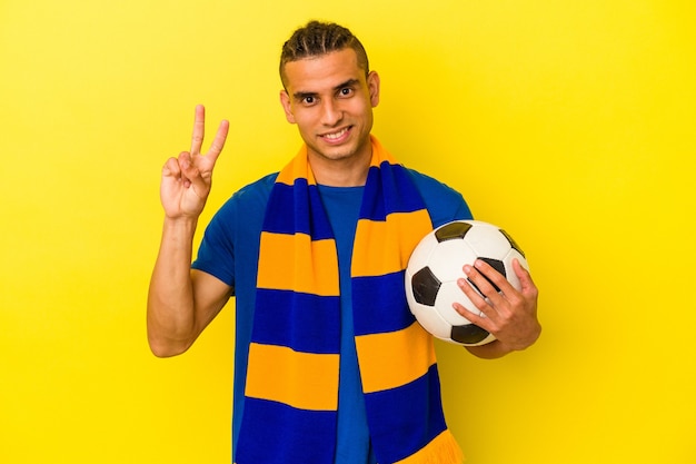 Jovem venezuelano assistindo futebol isolado em fundo amarelo, mostrando o número dois com os dedos.