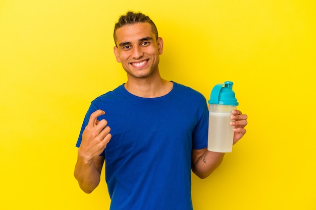 Jovem venezuelano bebendo um shake de proteína isolado em um fundo amarelo apontando com o dedo para você como se fosse um convite para se aproximar.