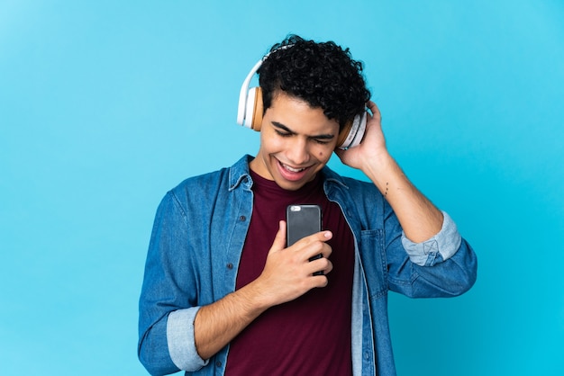 Jovem venezuelano isolado no azul ouvindo música com um celular e cantando