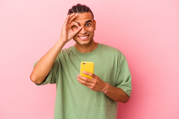 Jovem venezuelano segurando um telefone celular isolado no fundo rosa animado, mantendo o gesto ok no olho.