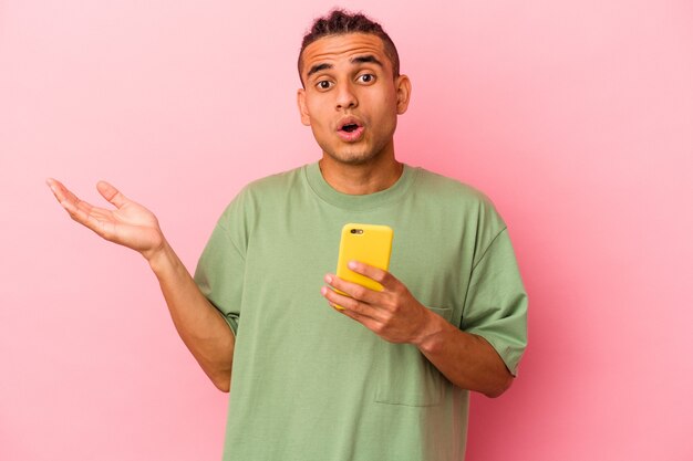 Jovem venezuelano segurando um telefone celular isolado no fundo rosa surpreso e chocado.
