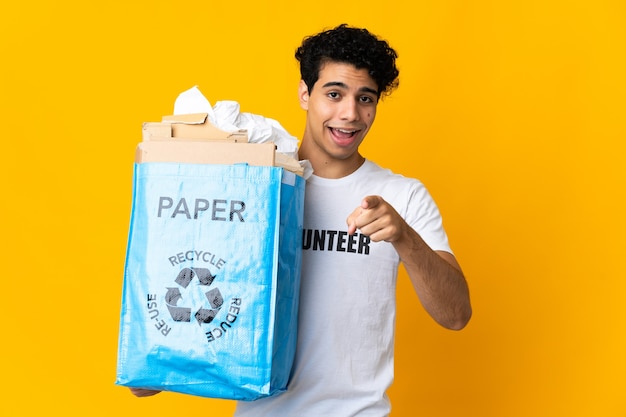 Jovem venezuelano segurando uma sacola de reciclagem cheia de papel para reciclar surpreso e apontando para a frente