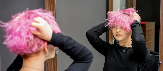 Jovem verificando seu novo penteado rosa curto encaracolado com as mãos na frente do espelho no salão de cabeleireiro