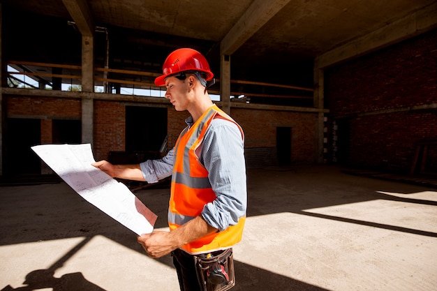 Jovem vestido com camisa, colete laranja e capacete explora a documentação da construção no canteiro de obras dentro do prédio em construção.