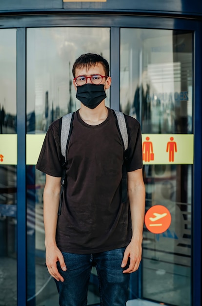 Jovem vestido de camiseta preta, calça jeans, máscara cirúrgica, óculos e mochila em frente a portas do aeroporto e olhando para a câmera