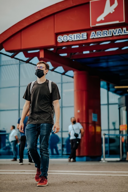 Jovem vestido de camiseta preta, calça jeans, tênis vermelho, mochila, máscara cirúrgica e óculos andando longe do aeroporto