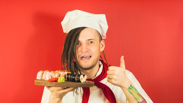 Jovem vestido de chef segurando a placa de madeira com rolos de sushi e mostrando gesto de como cozinheiro masculino com rolos de sushi apetitosos e mostrando o polegar sobre fundo vermelho