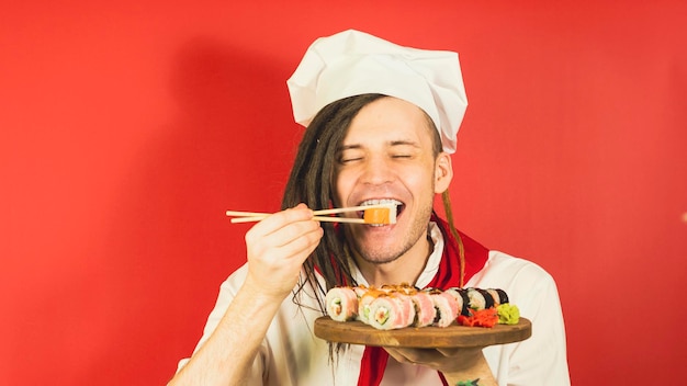 Jovem vestido de chef segurando a placa de madeira com rolos de sushi em fundo vermelho Cozinheiro masculino fechou os olhos comendo rolo de sushi apetitoso com pauzinhos