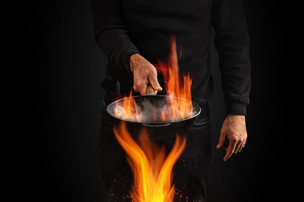 Foto jovem vestido de jumper e jeans, ele está segurando uma frigideira wok com fumaça acima do fogo contra bl ...