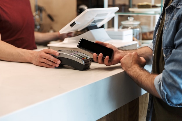 jovem vestindo camisa jeans pagando cartão de débito em um café enquanto garçom segurando um terminal de pagamento