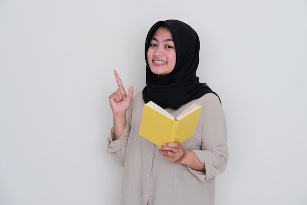 Jovem vestindo hijab sorrindo feliz e apontando para cima enquanto segura um livro