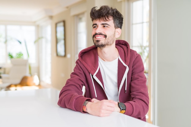 Jovem vestindo moletom casual sentado na mesa branca olhando para o lado com sorriso no rosto expressão natural Rindo confiante