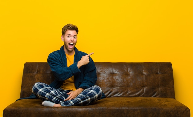 Jovem vestindo pijama olhando animado e surpreso apontando para o lado e para cima para copiar o espaço. sentado em um sofá