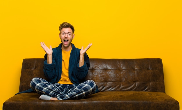 Jovem vestindo pijama, olhando feliz e animado, chocado com uma surpresa inesperada com as duas mãos abertas ao lado do rosto. sentado em um sofá