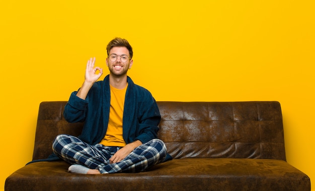 Jovem vestindo pijama, sentindo-se bem-sucedido e satisfeito, sorrindo com a boca aberta, fazendo sinal bem com a mão. sentado em um sofá