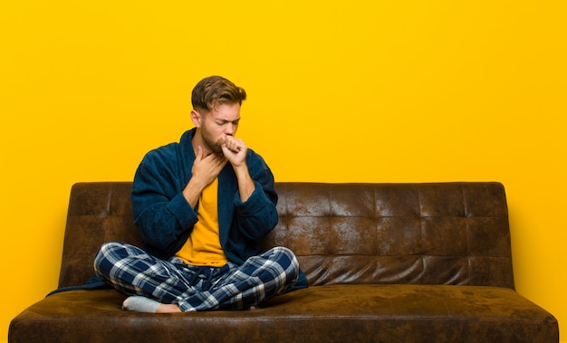 Jovem vestindo pijama, sentindo-se doente com uma dor de garganta e sintomas de gripe, tosse com a boca coberta. sentado em um sofá