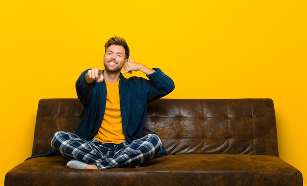 Foto jovem vestindo pijama sorrindo alegremente e apontando para a câmera enquanto fazia uma ligação, mais tarde, gesto, falando no telefone. sentado em um sofá